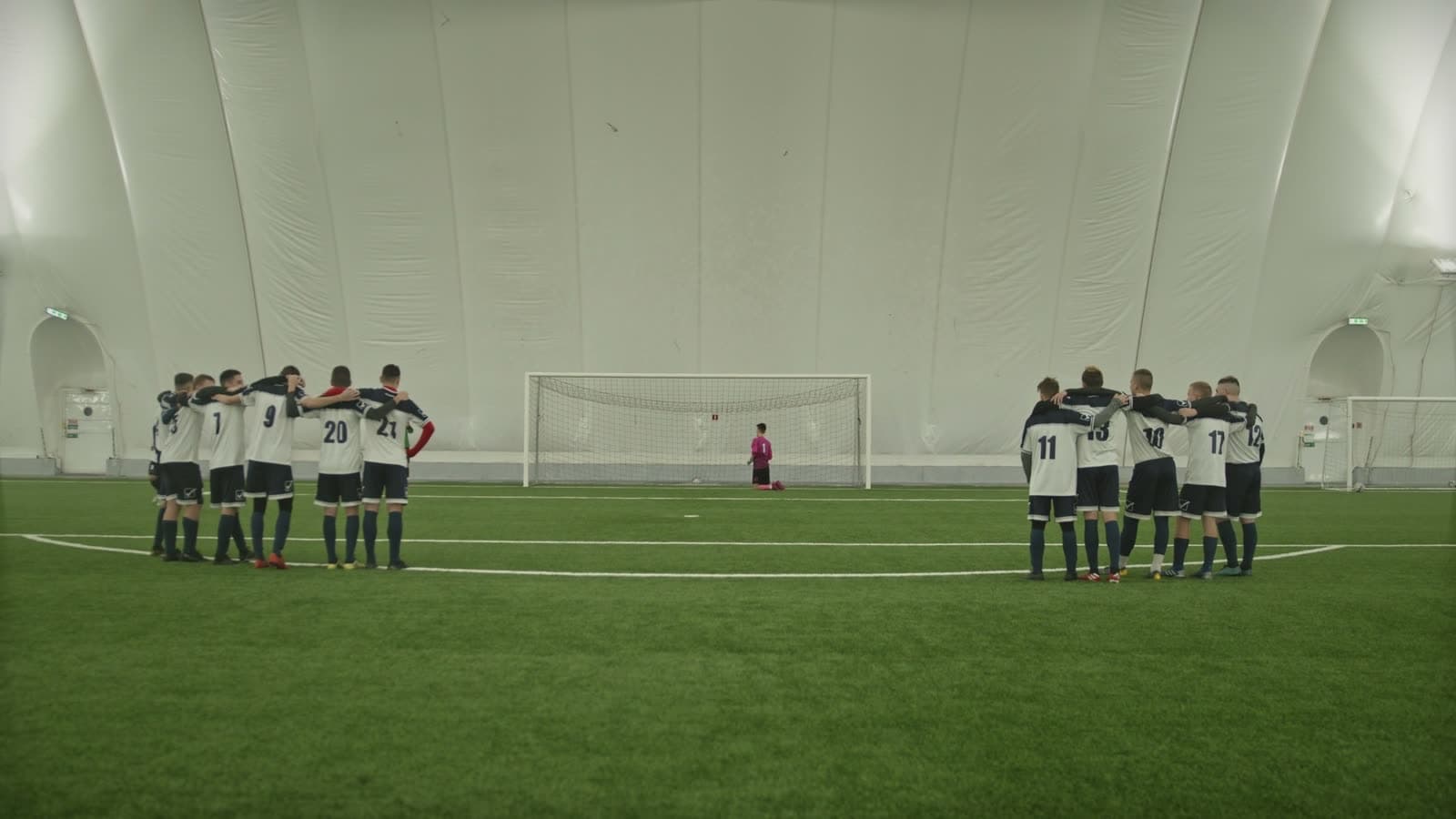 Two groups of indoor soccer players standing in team huddles with arms around each other on opposite sides of the pitch