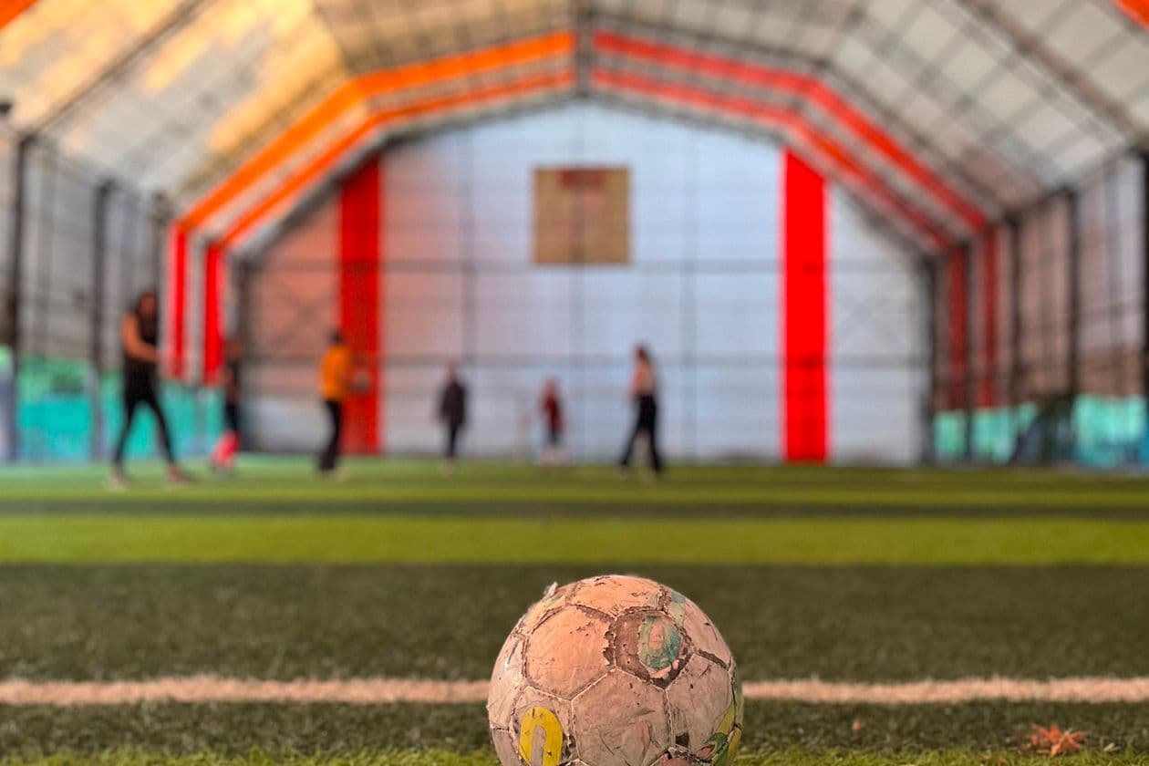 Indoor soccer players competing on artificial turf with a worn soccer ball in the foreground during an active match
