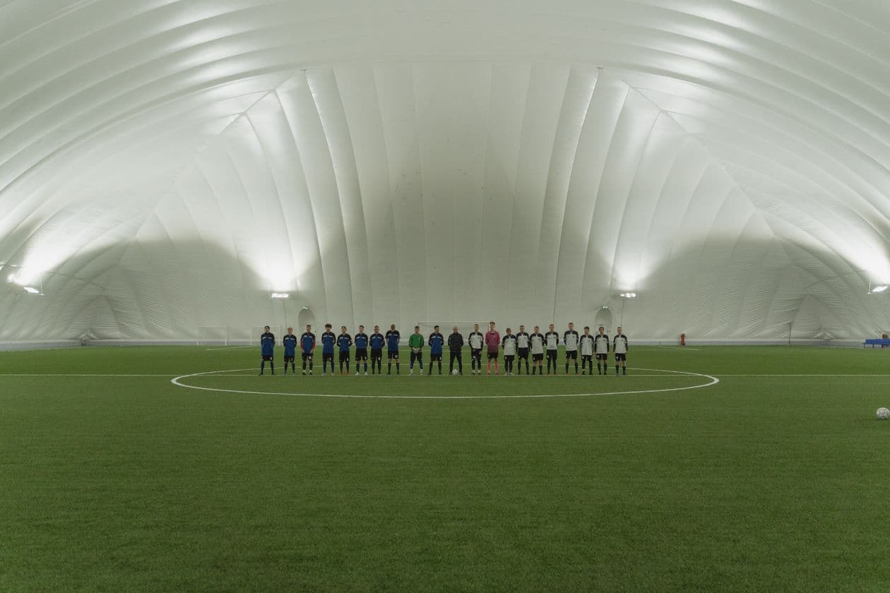 Two soccer teams lined up on an indoor artificial turf field under bright dome lighting before a match