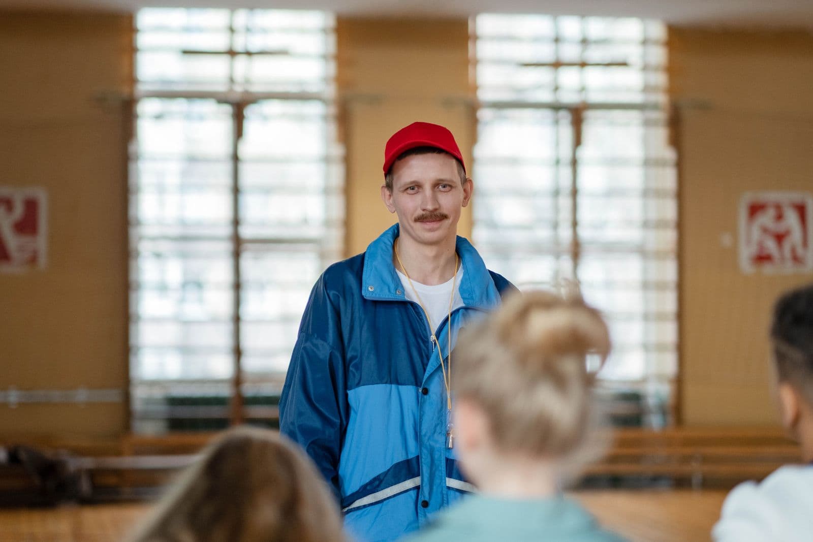 Male sports coach in a red cap and blue jacket addressing a group of young athletes inside an indoor sports hall