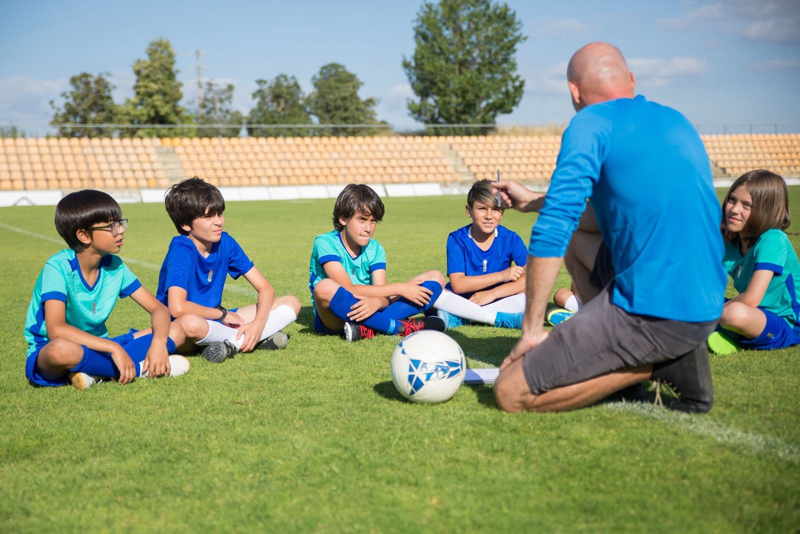 Sports coach sitting on the ground talking to a group of young soccer players during a training session on a grass pitch