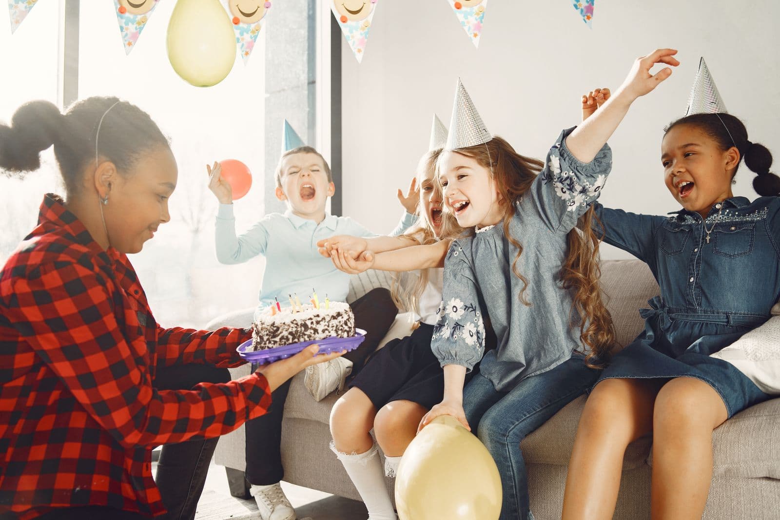Group of excited children in birthday party hats celebrating with arms raised and a birthday cake