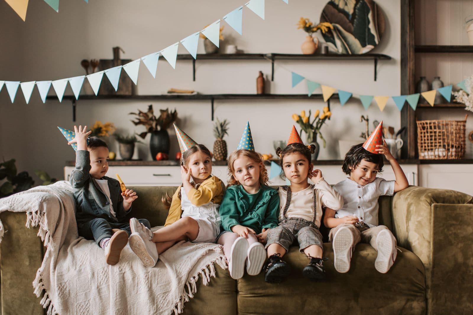 Five children wearing party hats sitting on a couch with bunting decorations at a birthday party